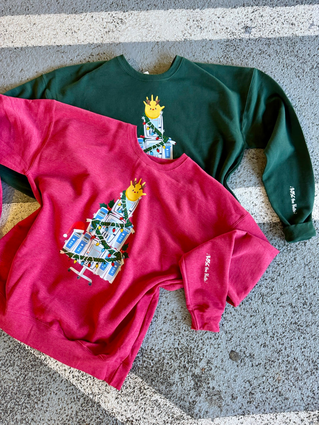 Two sweatshirts, one red and one green, with a IV Pump Christmas tree design and ROSC the Halls sleeve art, both on a concrete surface