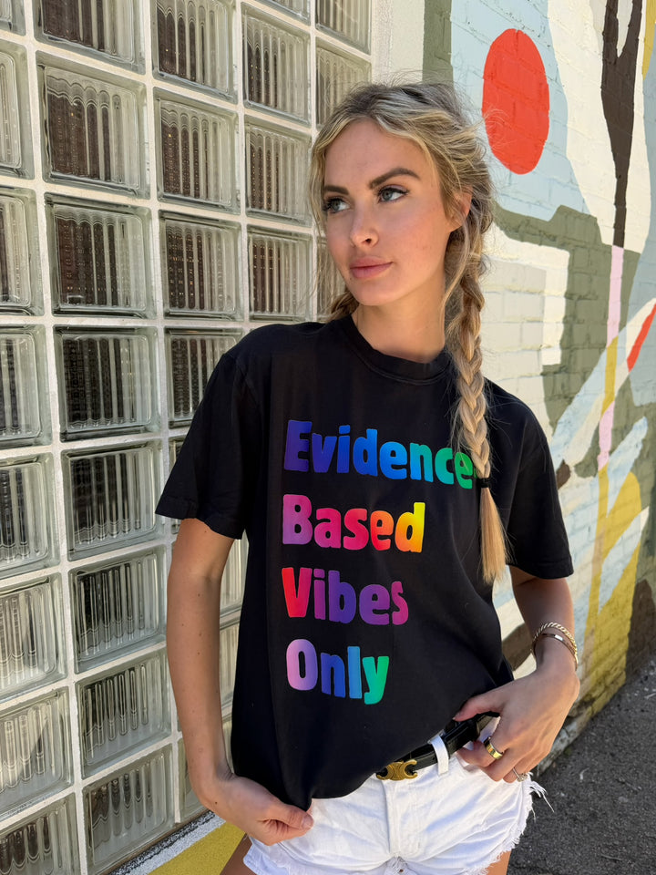 Person wearing a black t-shirt with colorful Evidence-Based Vibes Only text in front of a glass block wall and mural.