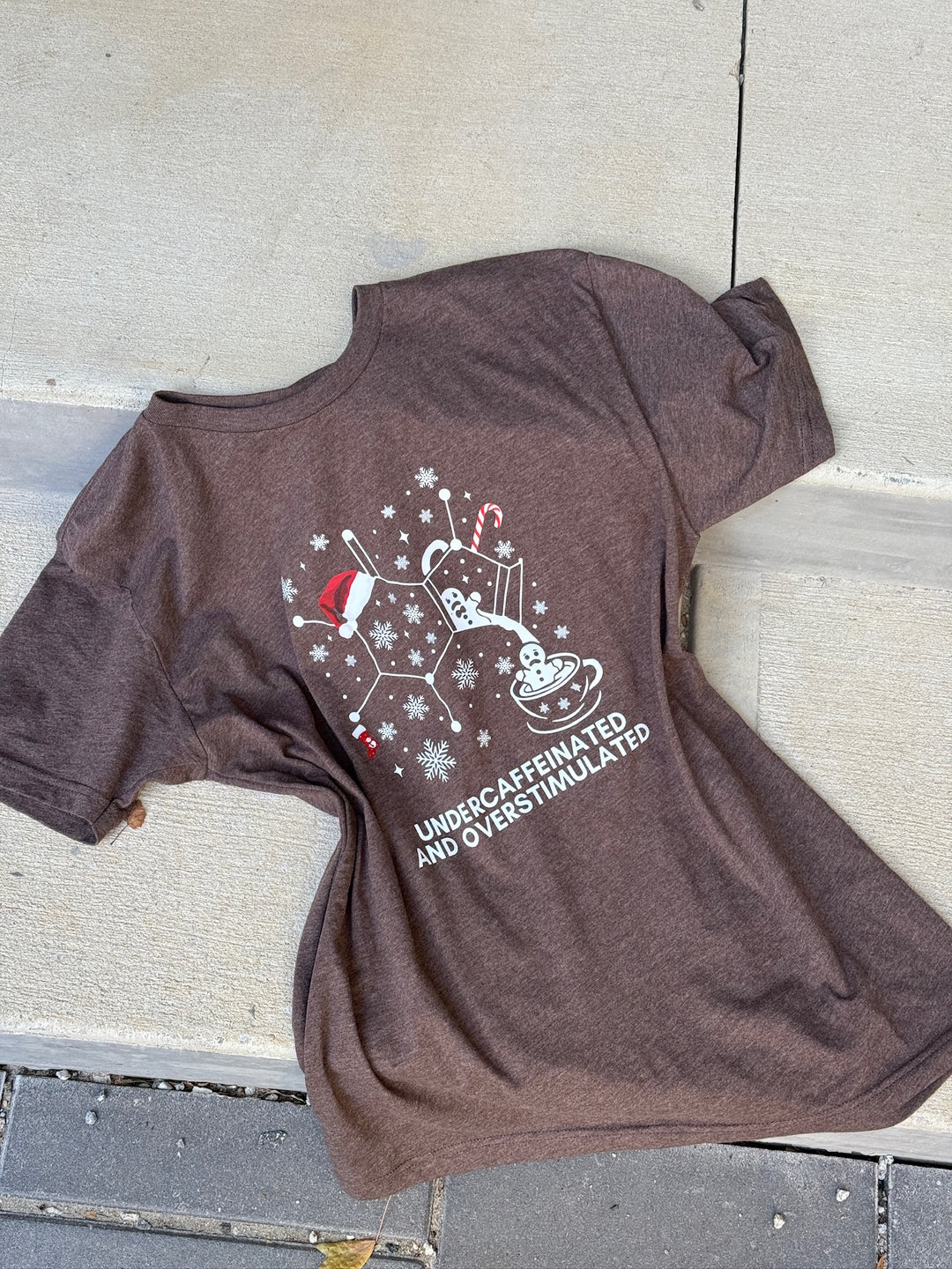 Brown t-shirt with a caffeine molecule coffee carafe Christmas design and 'undercaffeinated and overstimulated' text on a concrete surface