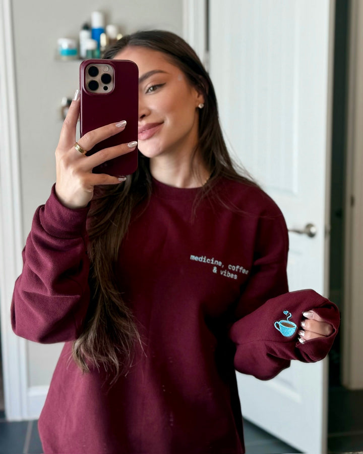 Dr Daisy wearing the maroon Medicine, Coffee & Vibes sweatshirt with a coffee cup design, taking a mirror selfie.