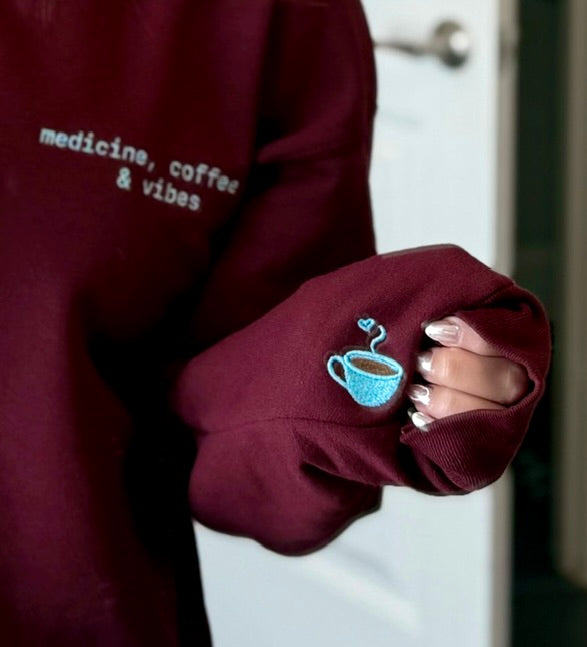 Maroon medicine, coffee and vibe sweatshirt with embroidered text and coffee cup design with steam heart detail, worn by Dr. Daisy.