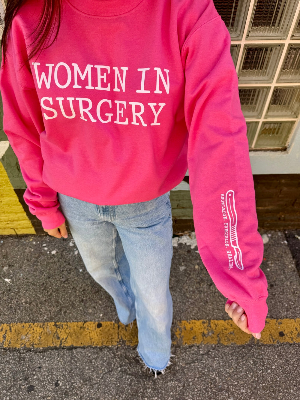 Person wearing a pink sweatshirt with 'WOMEN IN SURGERY' text, standing on a street.