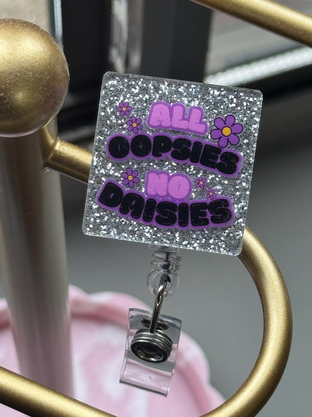 Retractable nurse badge reel with silver glittery background and bold text: “All Oopsies No Daisies” in purple and black with flower accents clipped on gold stand.