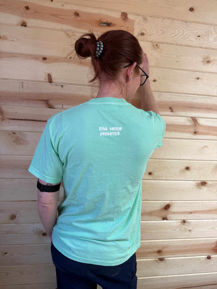 The back view of Ladyspinedoc (Dr. Betsy Grunch) wearing a light green T-shirt, showing the phrase "FINE MOTOR PROMOTER" printed in small white letters near the top of the shirt.