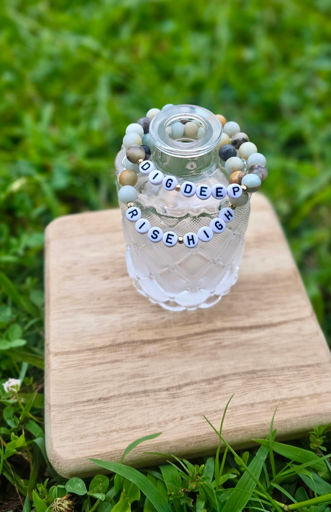Decorative bottle with 'DIG DEEP RISE HIGH' handmade bracelets on a wooden block outdoors.