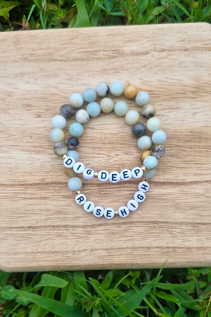 Two beaded bracelets with motivational Dig Deep Rise High on a wooden surface.