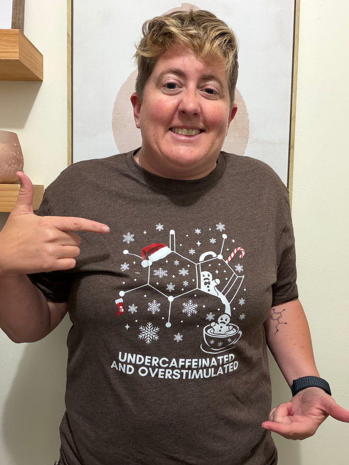 Dr. Beachgem wearing a brown holiday t-shirt with a graphic caffeine molecule design and text, pointing at the shirt.