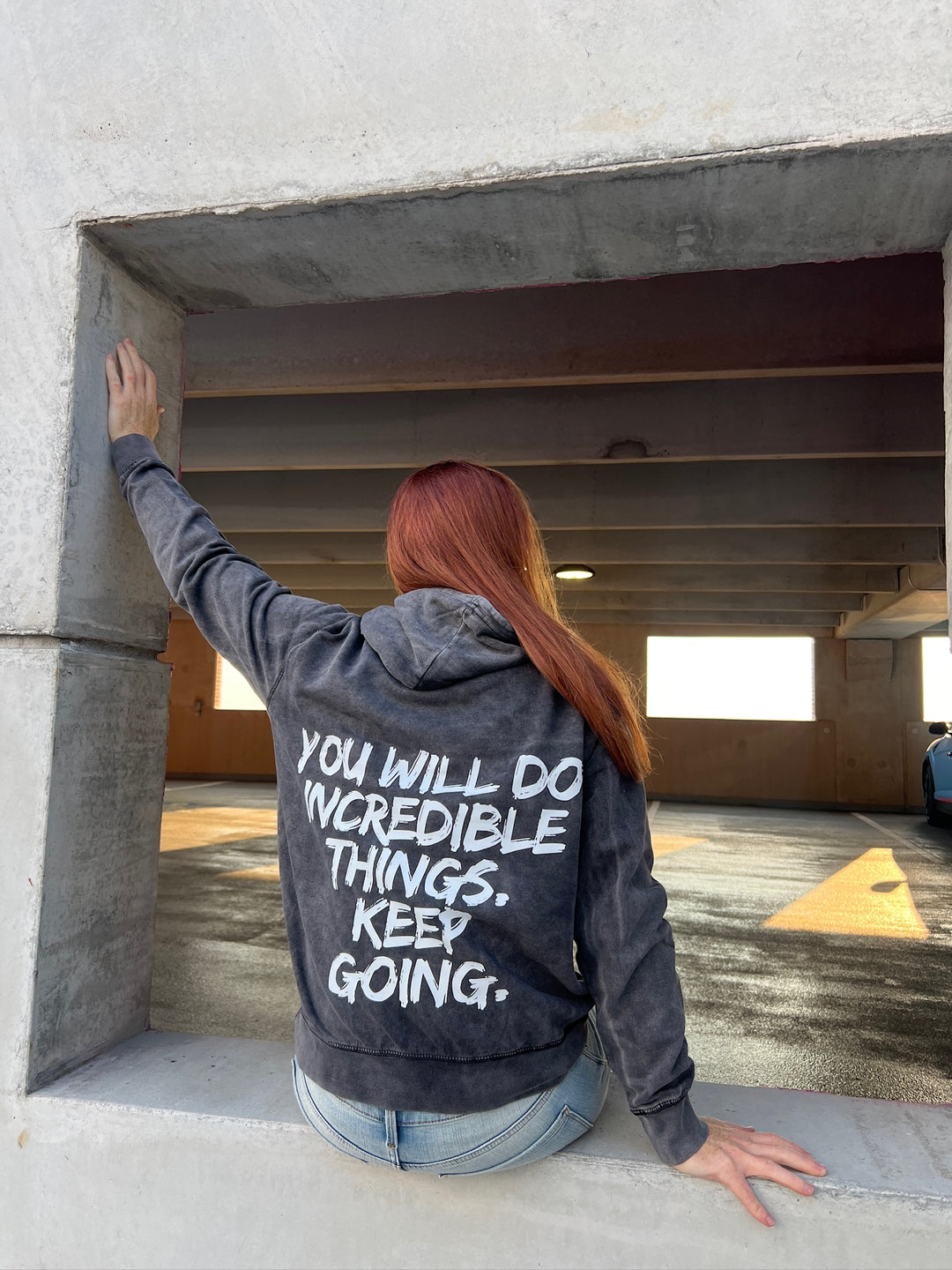  Dr. Betsy Grunch (Ladyspinedoc) sitting on a wall, displaying back of hoodie showing bold white brushstroke message: “YOU WILL DO INCREDIBLE THINGS. KEEP GOING.”
