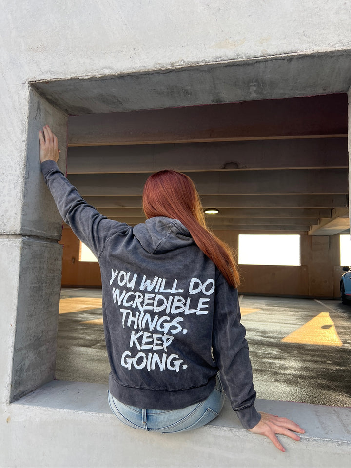  Dr. Betsy Grunch (Ladyspinedoc) sitting on a wall, displaying back of hoodie showing bold white brushstroke message: “YOU WILL DO INCREDIBLE THINGS. KEEP GOING.”
