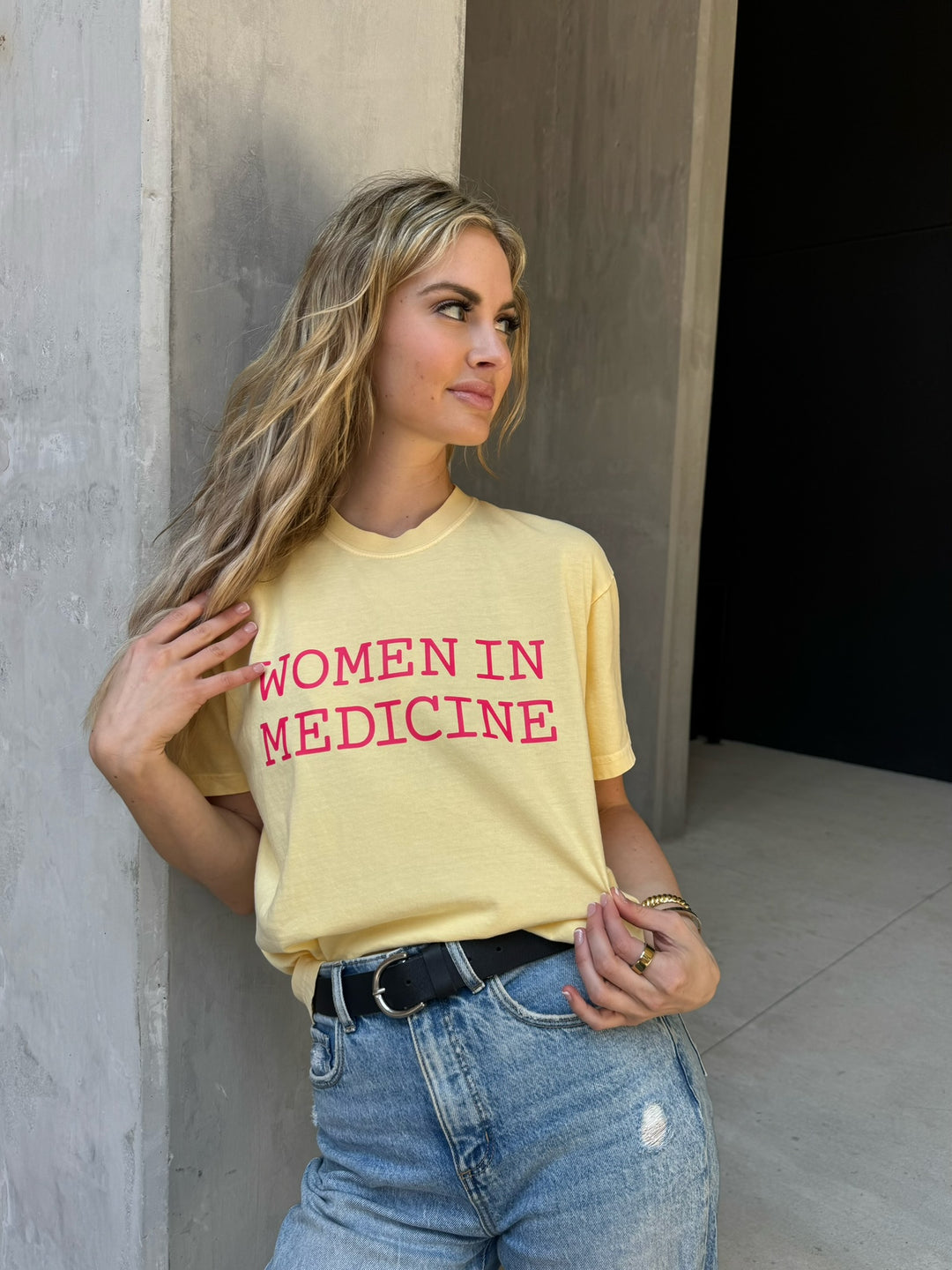 Woman wearing a yellow t-shirt with 'Women in Medicine' text, standing against a concrete wall.