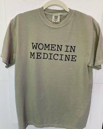 Green t-shirt with 'WOMEN IN MEDICINE' text on a hanger against a white wall.