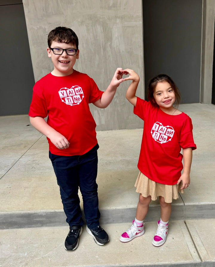 Two children wearing Atomic Attraction Tee Shirts - Kids Red Short Sleeve with Periodic Table Design that spells VALENTINE, and standing on a concrete surface.