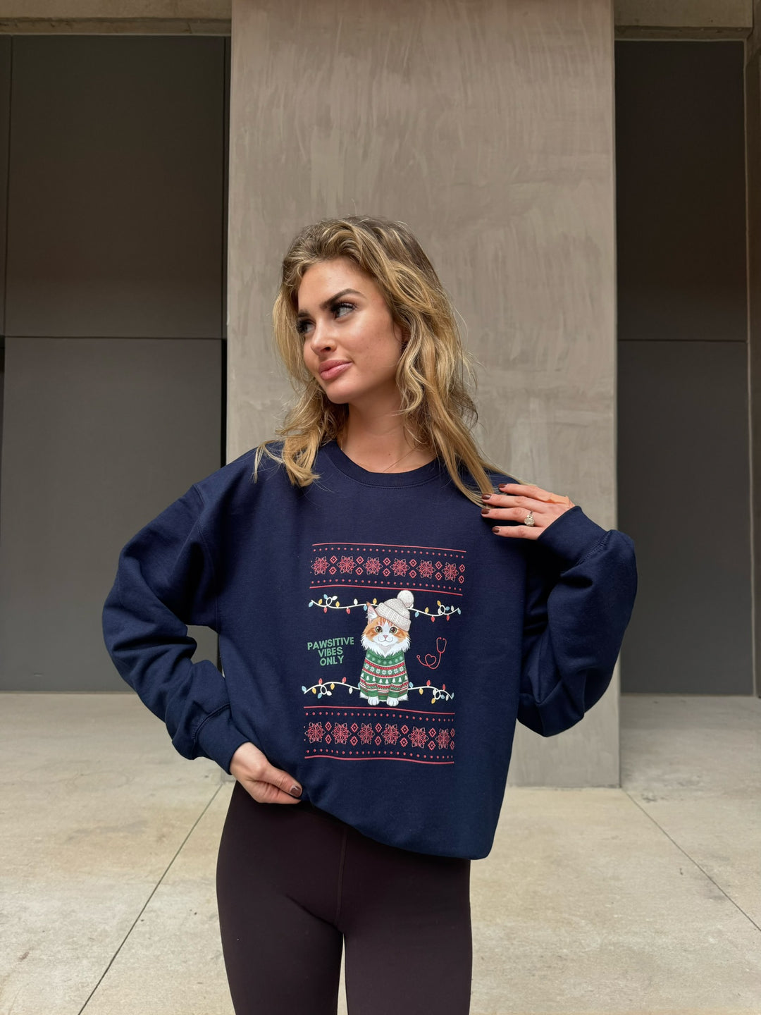 Woman wearing a navy blue shirt with a festive cat design, standing against a neutral wall.