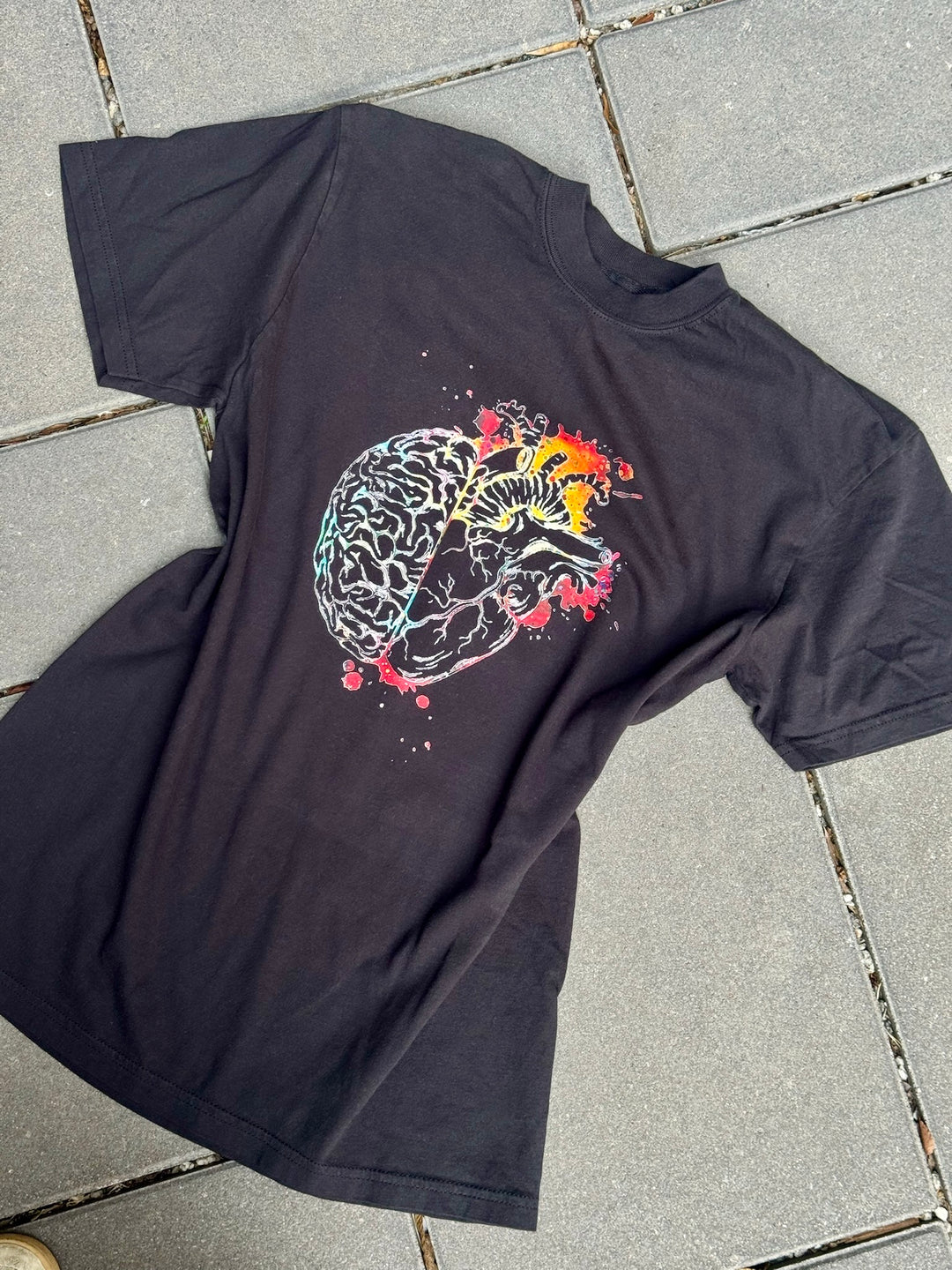 Black t-shirt with a colorful anatomical brain/heart design on a tiled floor