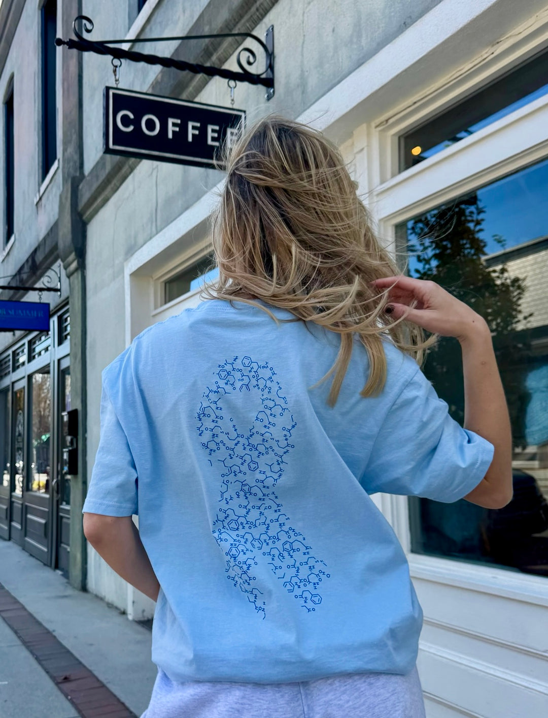 Woman wearing a light blue t-shirt with a ribbon design on the back made up of insulin molecules for diabetes awareness, standing in front of a coffee shop.