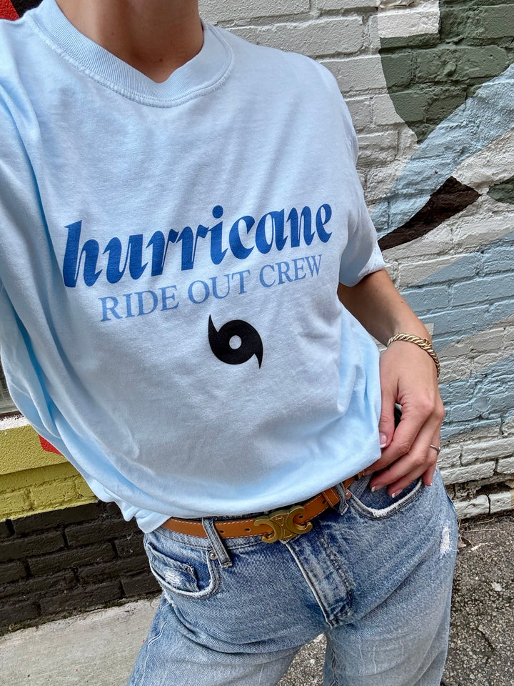 Person wearing a light blue t-shirt with 'hurricane RIDE OUT CREW' text and logo, standing against a stone wall.