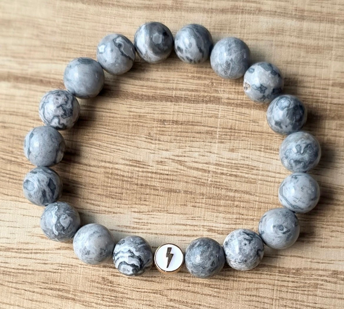 Flat lay of Glioblastoma Awareness Bracelet made of gray Map Stone Jasper beads with one white enamel bead with gold lightning bolt.