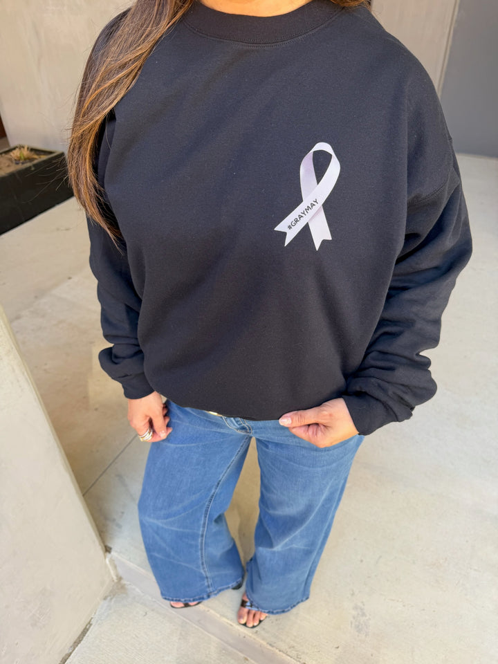 Person wearing a black sweatshirt with a gray ribbon logo on a neutral background. The ribbon says #graymay.