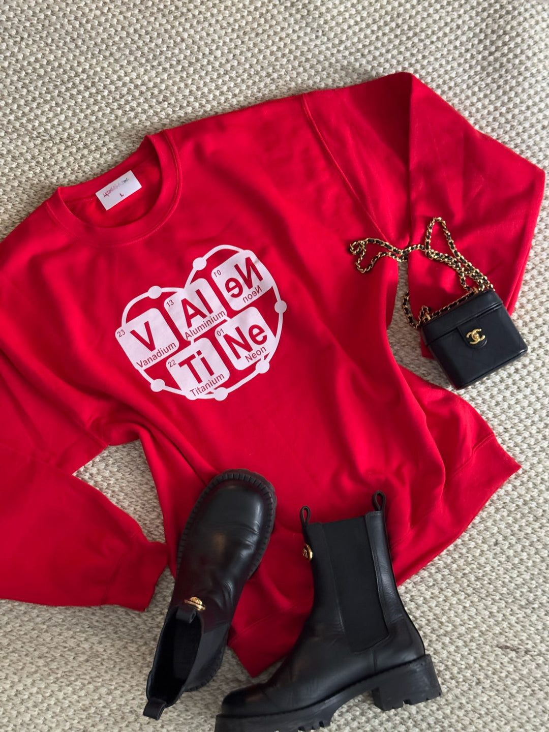 Front view of red Atomic Attraction crewneck sweatshirt featuring periodic table spelling “Valentine” with orbiting heart atom design on a sisal rug with black boots and a small black Chanel bag
