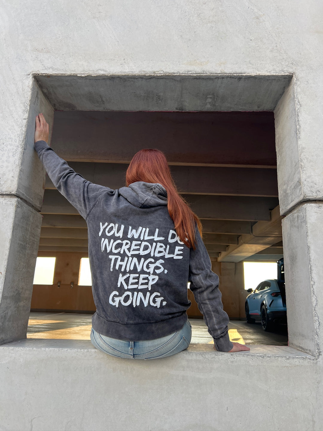 Dr. Betsy Grunch (Ladyspinedoc) sitting on a wall, facing away, displaying back of hoodie showing bold white brushstroke message: “YOU WILL DO INCREDIBLE THINGS. KEEP GOING.”
