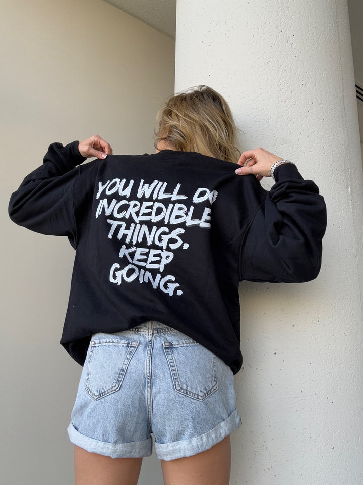 Model wearing The Incredible Sweatshirt, showing the back view of the shirt with bold white brushstroke message: "YOU WILL DO INCREDIBLE THINGS. KEEP GOING."
