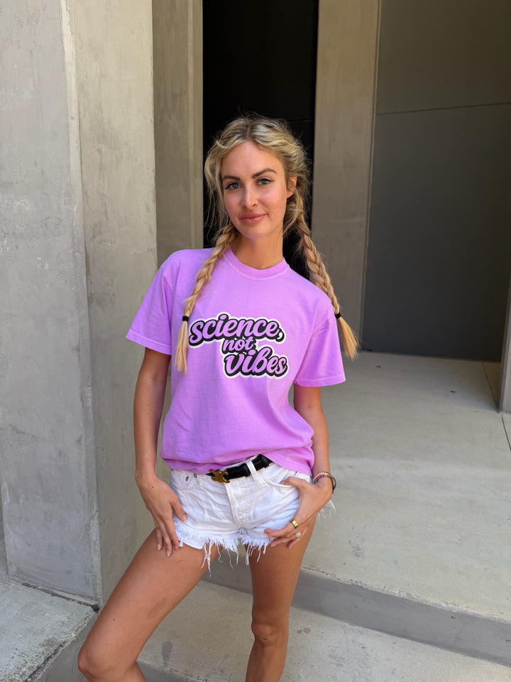 Woman wearing a purple t-shirt with 'Science Not Vibes' text, standing against a concrete wall.
