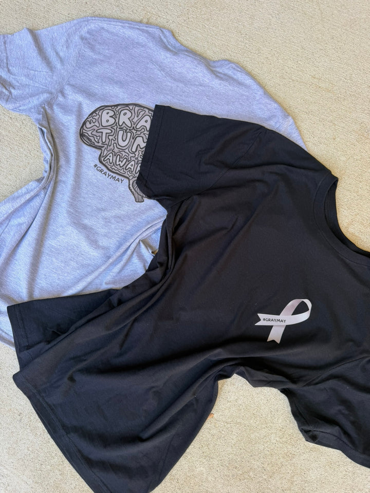 Gray shirt with brain graphic and black shirt with gray ribbon logo on a beige surface