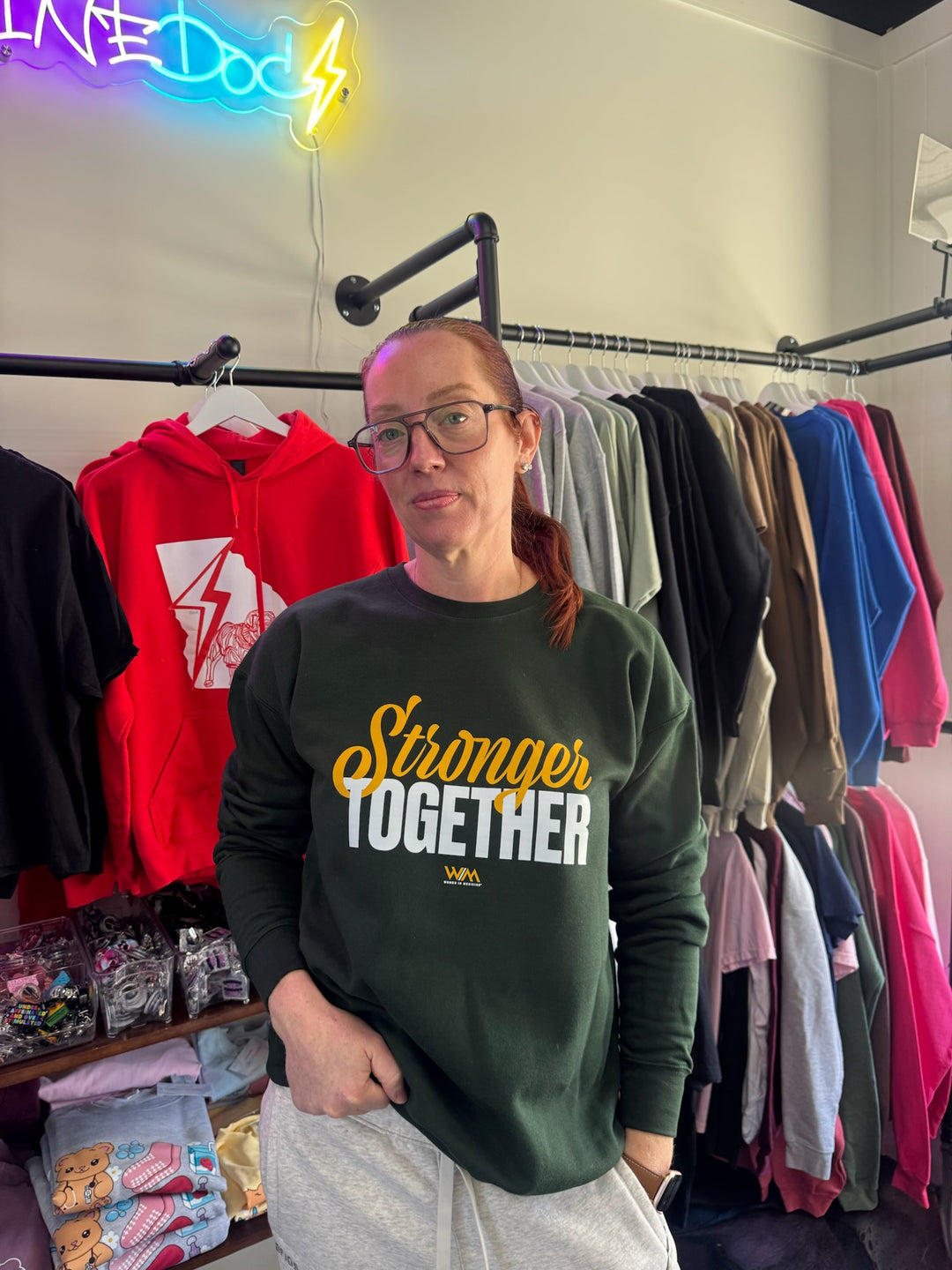 Ladyspinedoc wearing a green sweatshirt with 'Stronger Together' text in her clothing store.