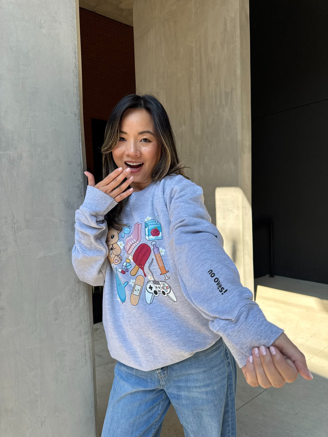 Model wearing Ladyspinedoc X Scrubhacks No Owies! Pediatrics Collage Crewneck Sweatshirt - Heather Gray With Pedi Nurse Essentials and showing off the “No Owies!” on Sleeve