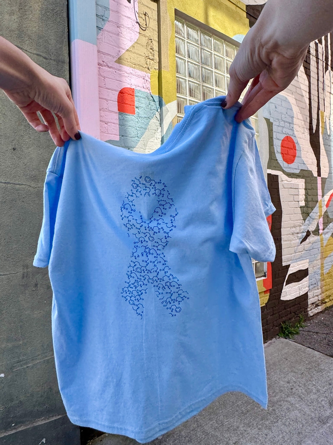 Back of light blue youth t-shirt with a blue ribbon design made up of insulin molecules, held by two hands against a colorful mural background.