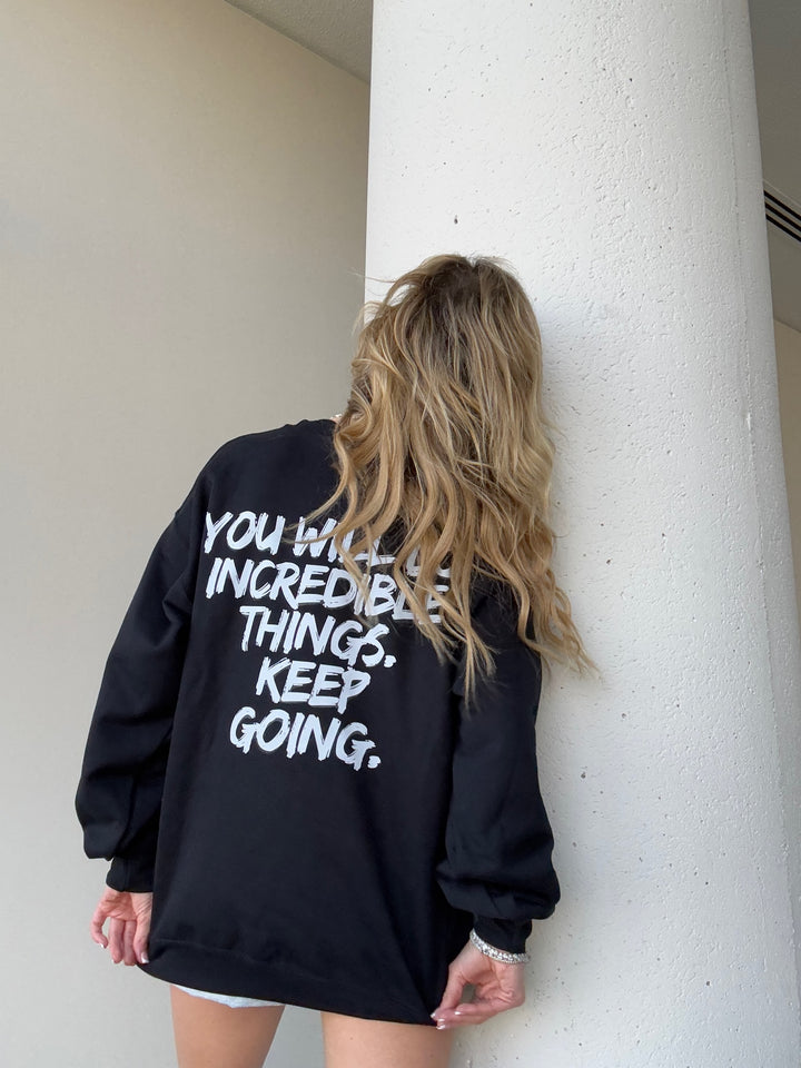 Model holding down the bottom hem and showing the back of the black Incredible Sweatshirt with bold white brushstroke message: "YOU WILL DO INCREDIBLE THINGS. KEEP GOING."
