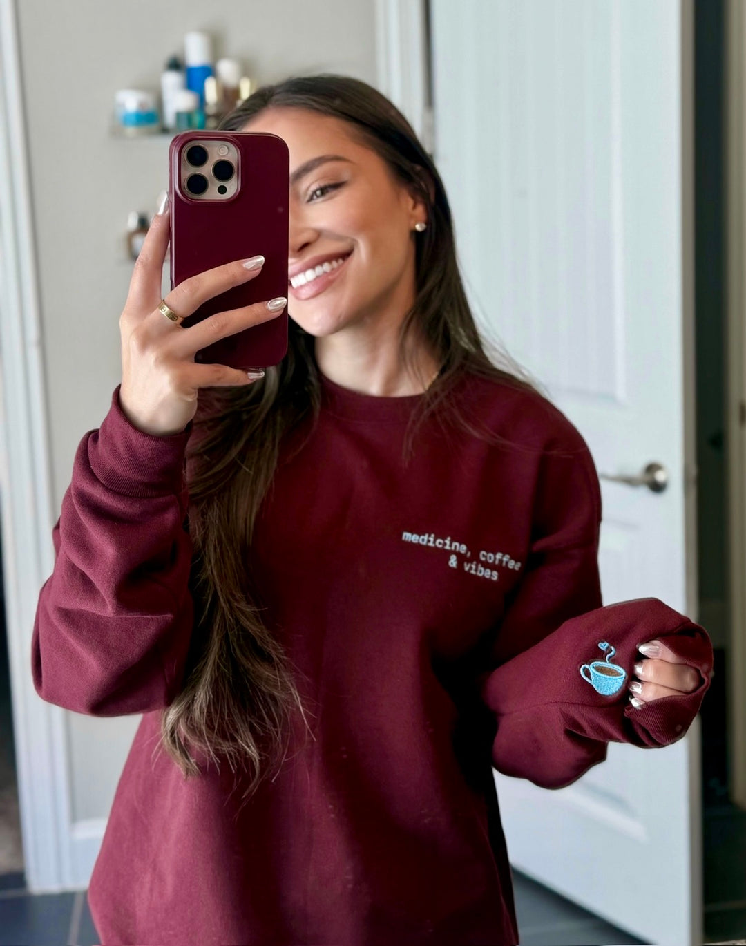 Dr Daisy wearing a maroon Medicine, Coffee & Vibes sweatshirt, showing off the coffee cup graphic on sleeve.