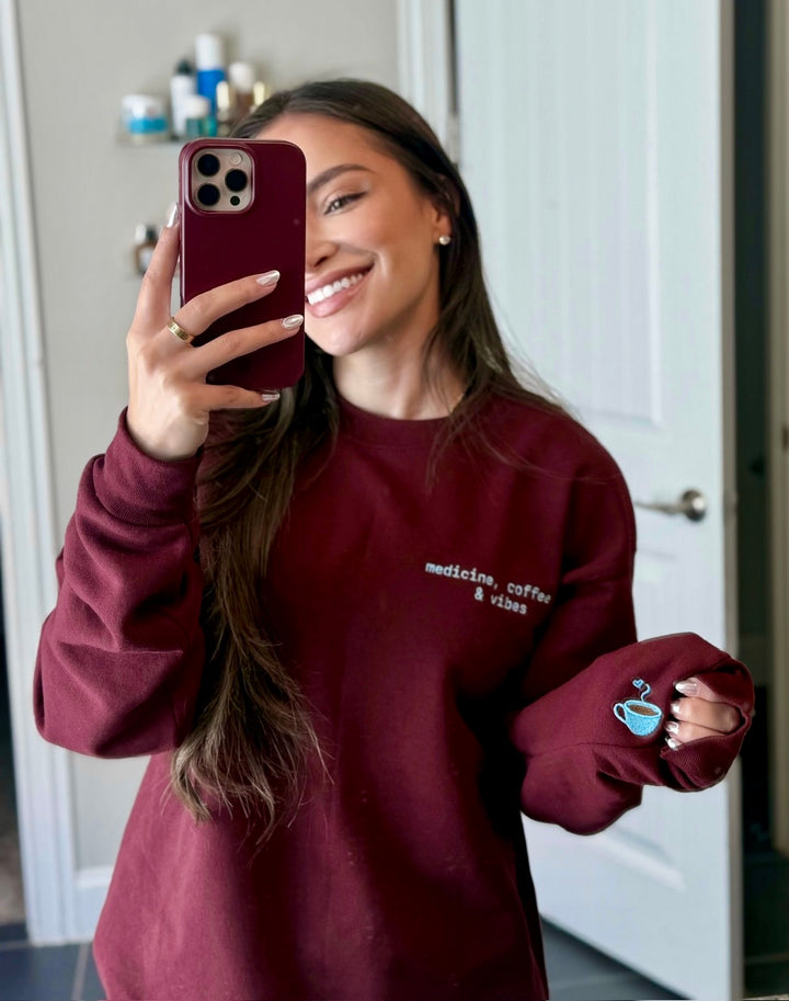 Dr Daisy wearing a maroon Medicine, Coffee & Vibes sweatshirt, showing off the coffee cup graphic on sleeve.