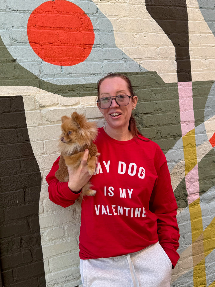 Ladyspinedoc wearing a red long-sleeve shirt with 'My dog is my Valentine' text, holding a small dog, standing in front of a colorful wall.