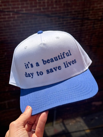 White and blue cap with embroidered "It's a beautiful day to save lives" text held against a brick wall