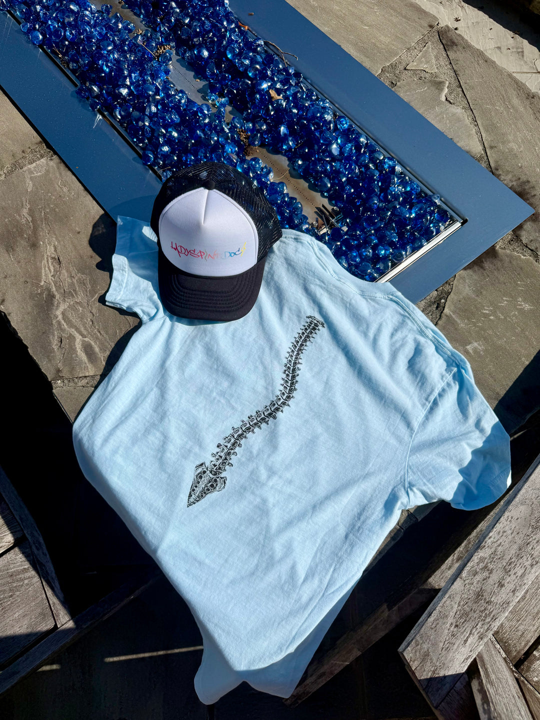 Flat lay showing the back of the Bent, Not Broken Scoliosis Awareness Tee - Blue Cotton T-Shirt with a Ladyspinedoc hat next to it.