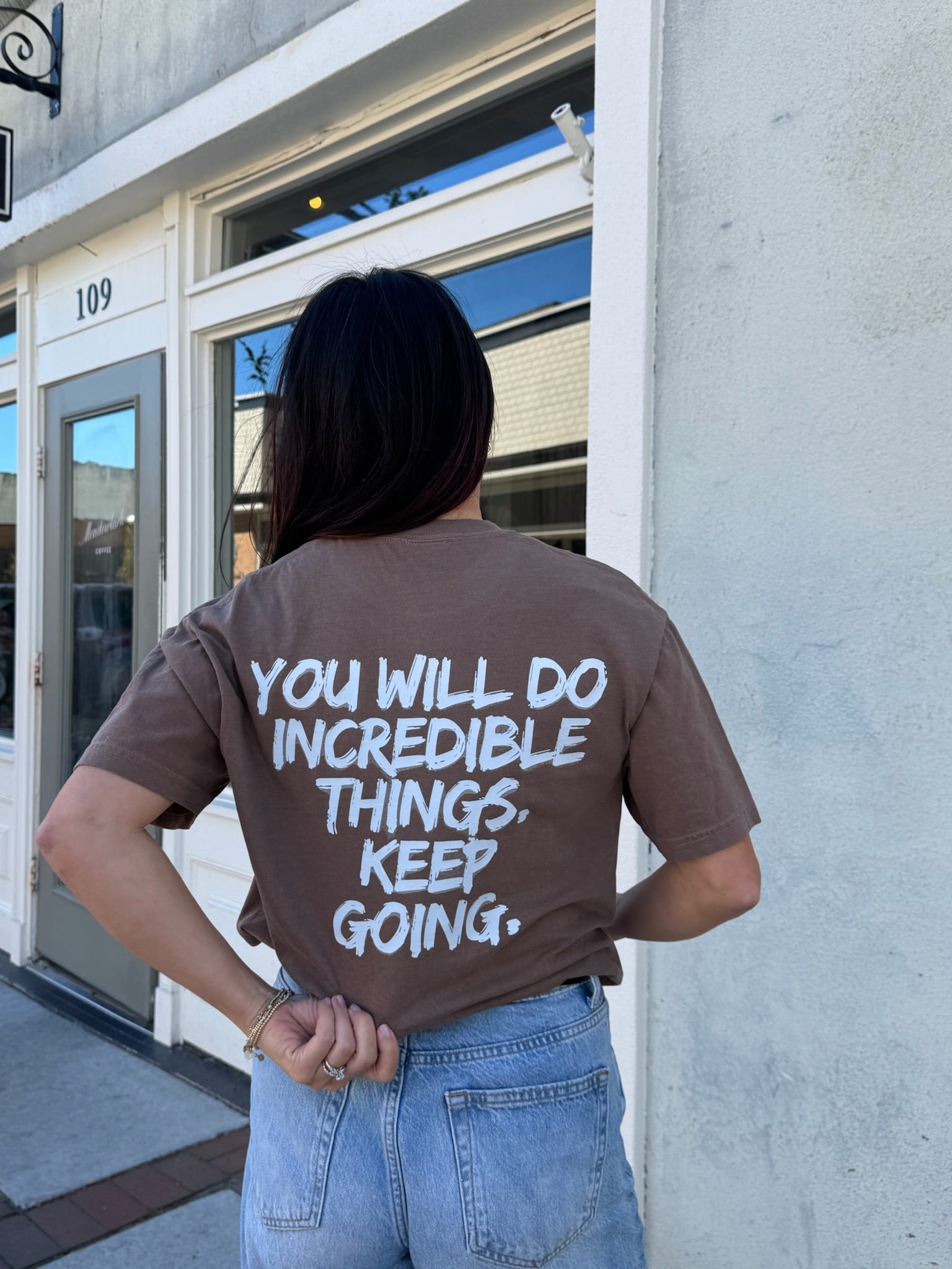 Person wearing The Incredible Tee Shirt - Brown Shirt with Motivational "YOU WILL DO INCREDIBLE THINGS. KEEP GOING." Motto, standing outside a building.