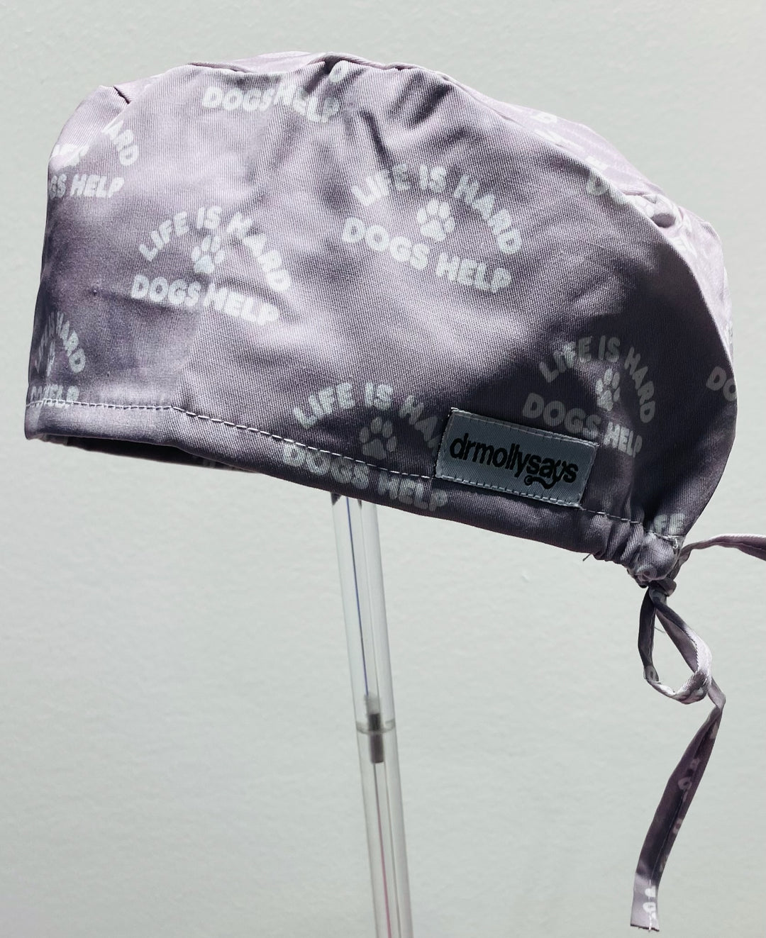 Gray scrub hat with 'Life is Hard, Dogs Help' text and a visible brand label on a light gray background.