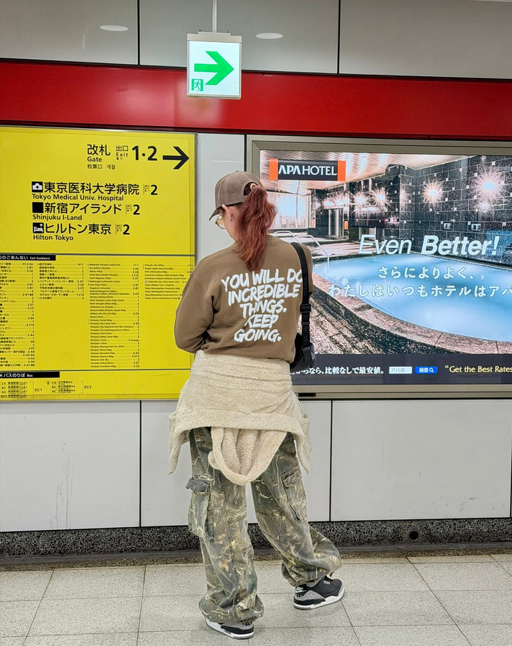Dr. Betsy Grunch wearing her Ladyspinedoc brown Incredible sweatshirt,  standing in a Japanese subway station looking at a large screen advertisement.