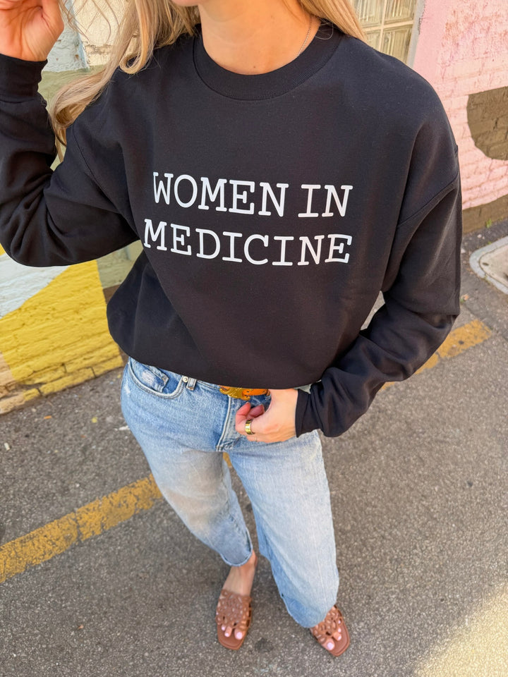 Person wearing a black sweatshirt with 'WOMEN IN MEDICINE' white text on a street.