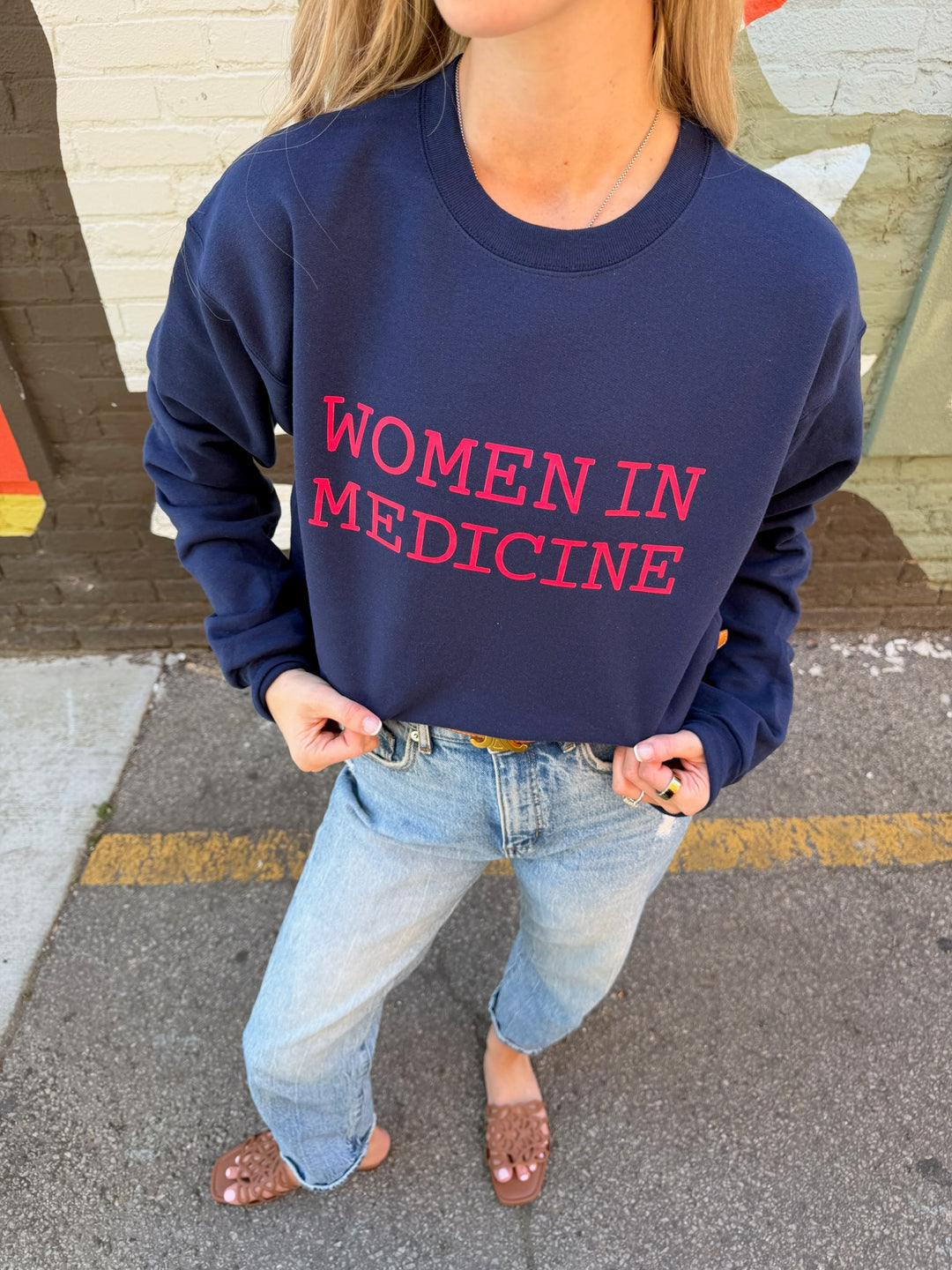 Person wearing a navy blue sweatshirt with 'WOMEN IN MEDICINE' in pink text, standing on a street.