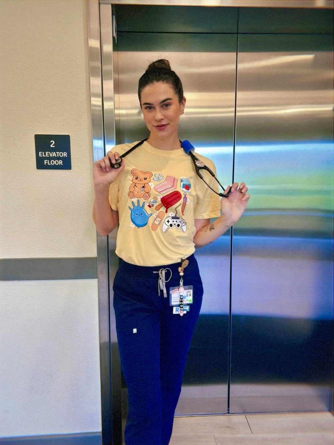 Scrubhacks holding a stethoscope and wearing yellow Pediatrics Collage Tee Shirt  in front of elevator