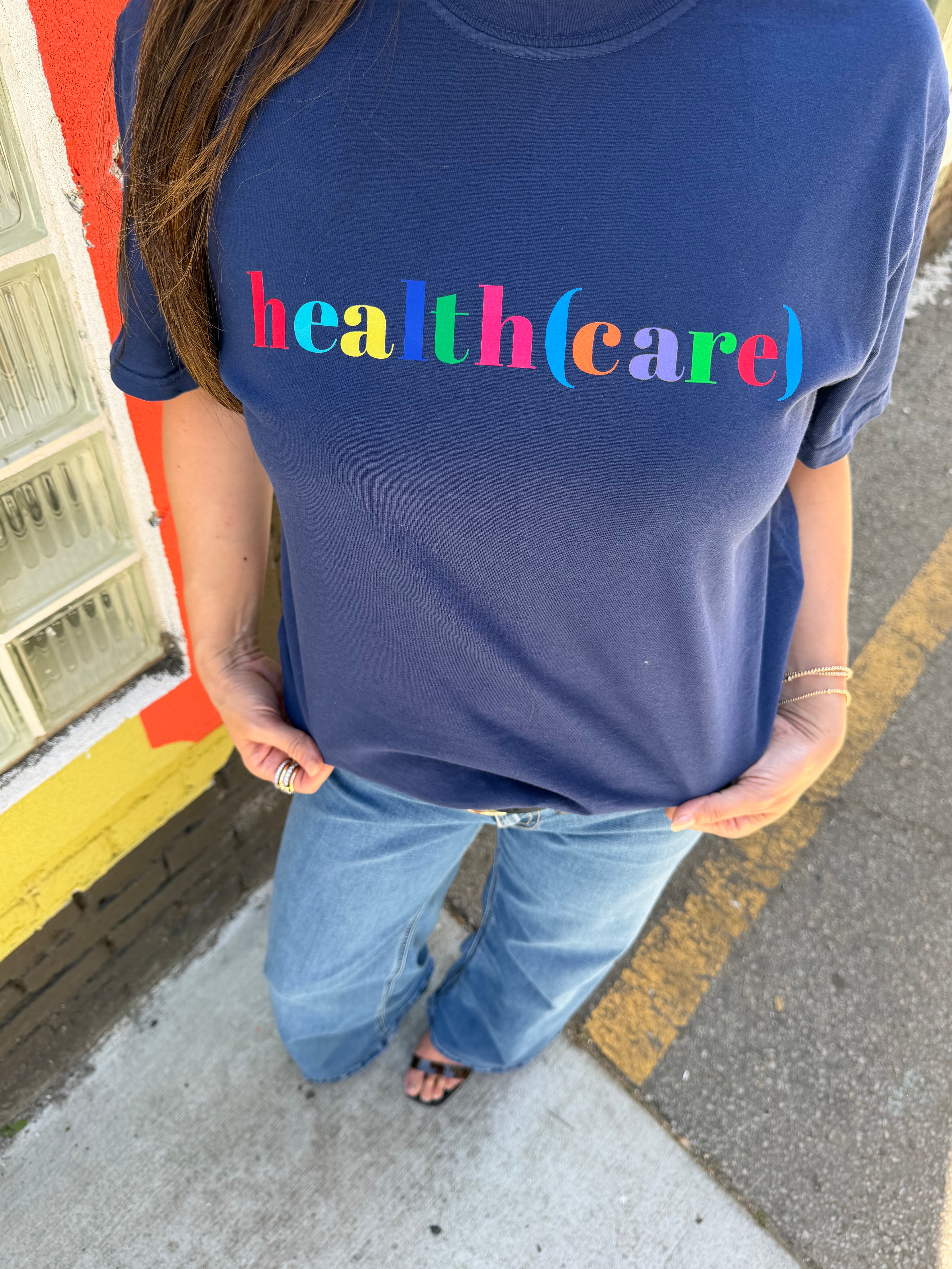 Person wearing a navy blue shirt with 'health(care)' printed on it, standing on a sidewalk.
