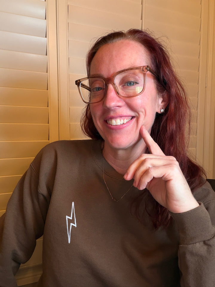 Ladyspinedoc wearing a brown Incredible sweatshirt with a lightning bolt logo, smiling and posing with hand on chin. 