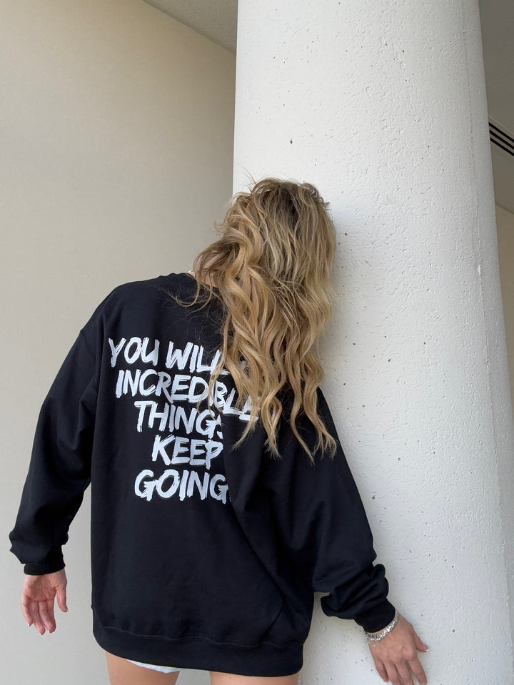 Model showing the back of the black Incredible Sweatshirt with bold white brushstroke message: "YOU WILL DO INCREDIBLE THINGS. KEEP GOING."

