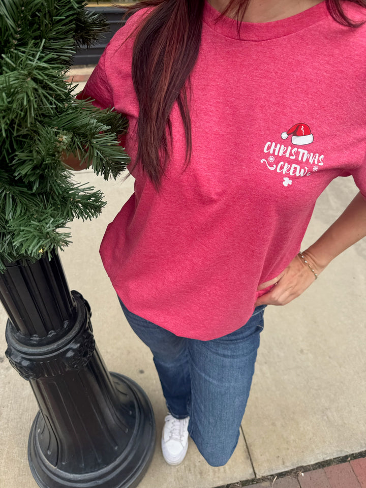 Person wearing a red t-shirt with a Christmas Crew design, standing next to a small tree.