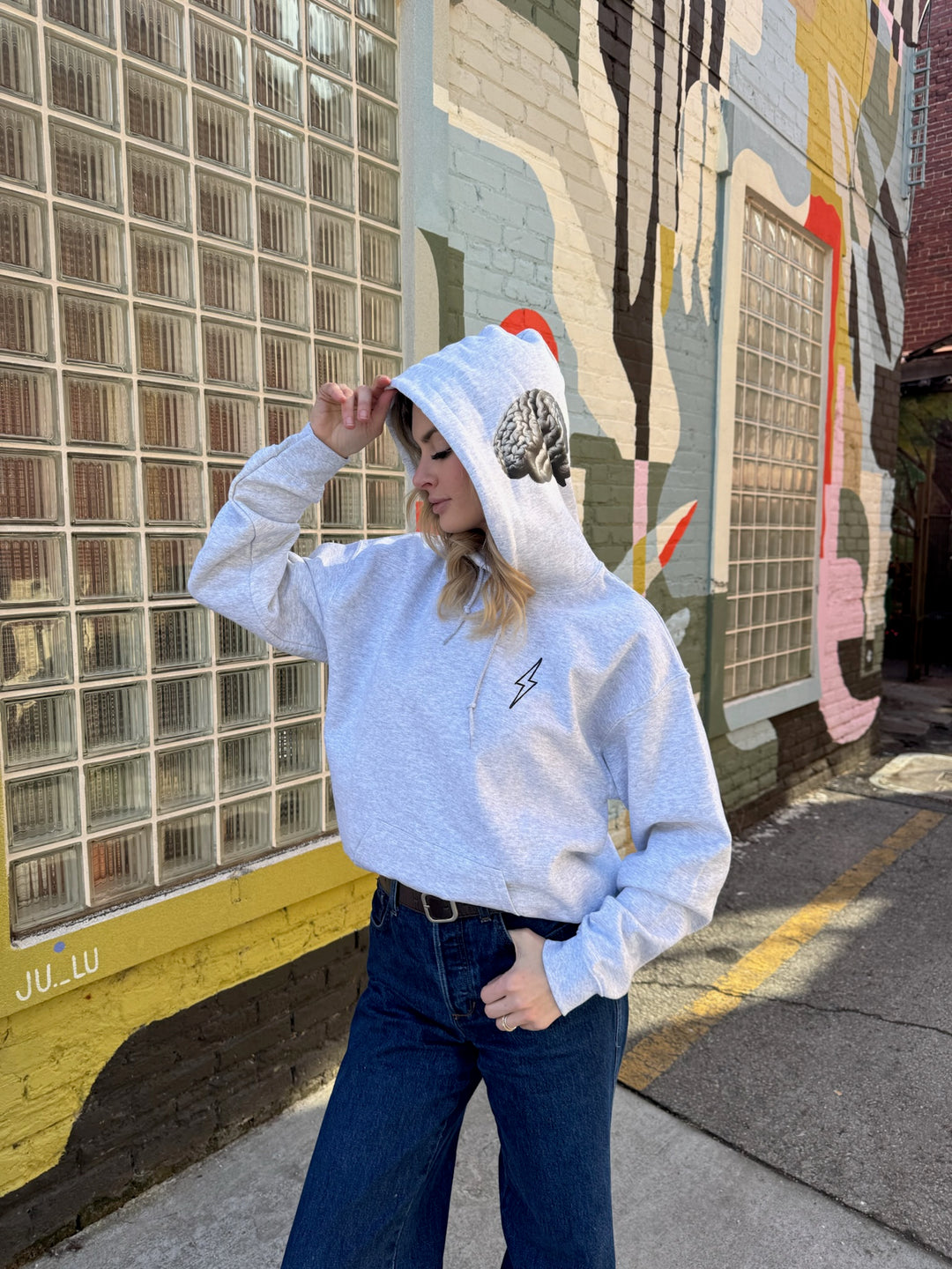 Person wearing The Cerebrospinal Hoodie - Light Gray Anatomical Brain and Spine Graphic Sweatshirt and dark jeans standing in front of a colorful mural.