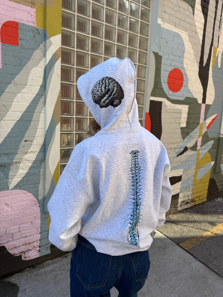 Person wearing The Cerebrospinal Hoodie - Light Gray Anatomical Brain and Spine Graphic Sweatshirt, standing in front of a colorful mural.