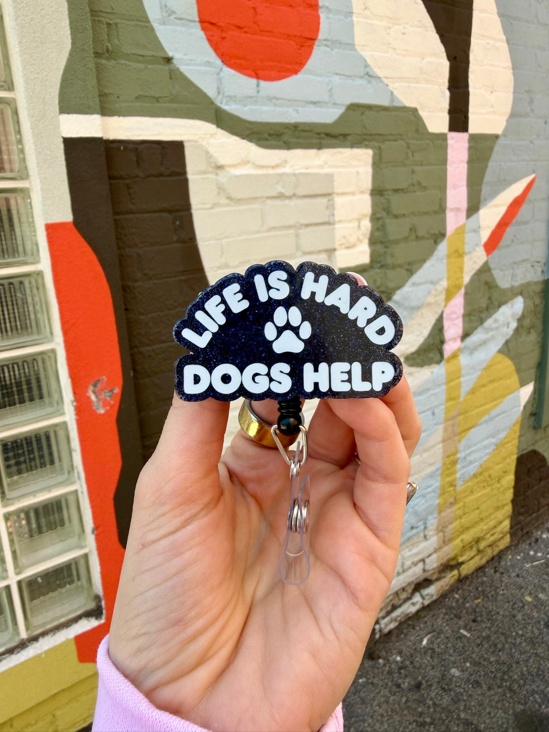 Badge holder with 'Life is Hard, Dogs Help' text held in front of a colorful mural.