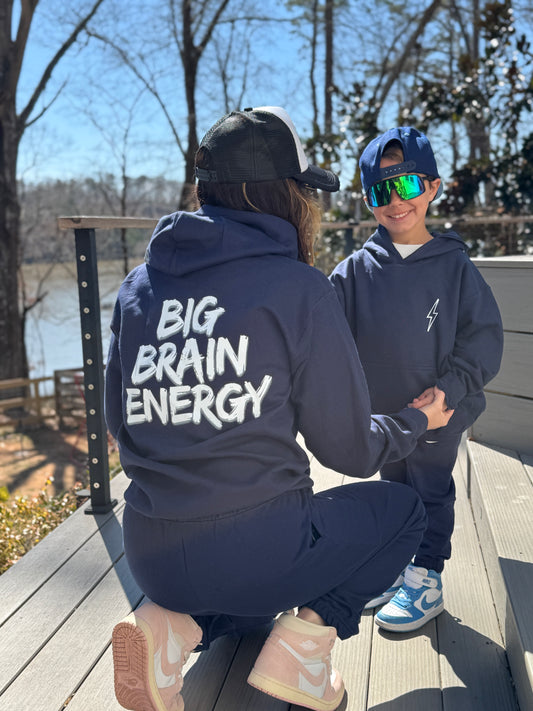 Adult model wearing Navy hoodie with "BIG BRAIN ENERGY" printed in bold white capital letters across the back. Child model showing front view of Big Brain Energy Youth Hoodie with lightning bolt on upper left chest and matching Big Brain Energy Youth Sweatpants.
